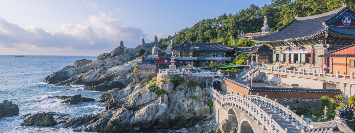 Korea-temple-by-the-sea-on-rocky-coastline-at-dusk, Traditional Korean temple architecture along the scenic coastline with ocean views and lush hillside, ideal for cultural tourism and spiritual retreats, SEO for Korea tourism and cultural heritage.