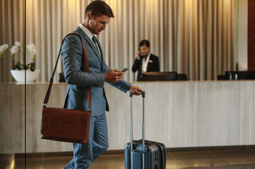 A professional man in a gray suit with a brown leather bag and rolling suitcase standing in an airport terminal, looking at his phone before travel.