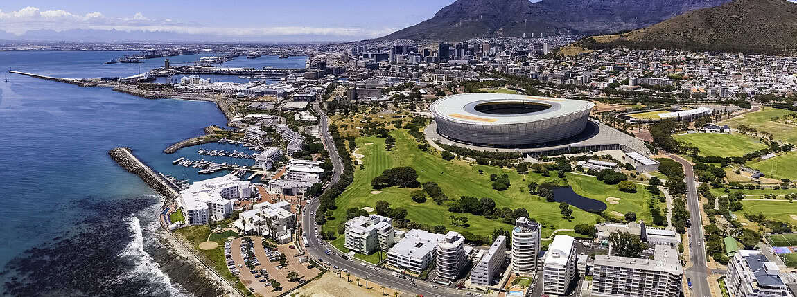 Aerial view of Cape Town's stadium, waterfront, and surrounding neighborhoods with Table Mountain in the background, showcasing urban development, sports facilities, and coastal scenery.