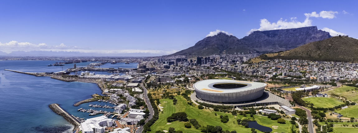 Aerial view of Cape Town's stadium, waterfront, and surrounding neighborhoods with Table Mountain in the background, showcasing urban development, sports facilities, and coastal scenery.