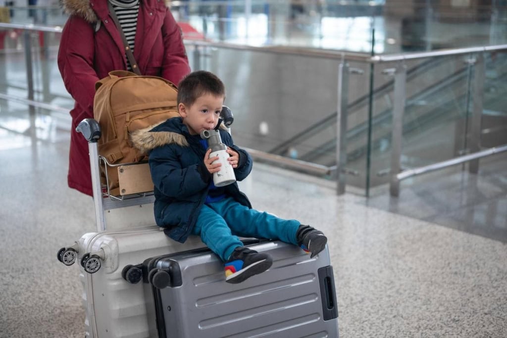 An adorable young boy in a winter jacket sits on a silver suitcase in an airport, drinking from a water bottle, with a backpack behind him, ready for travel or international journey.