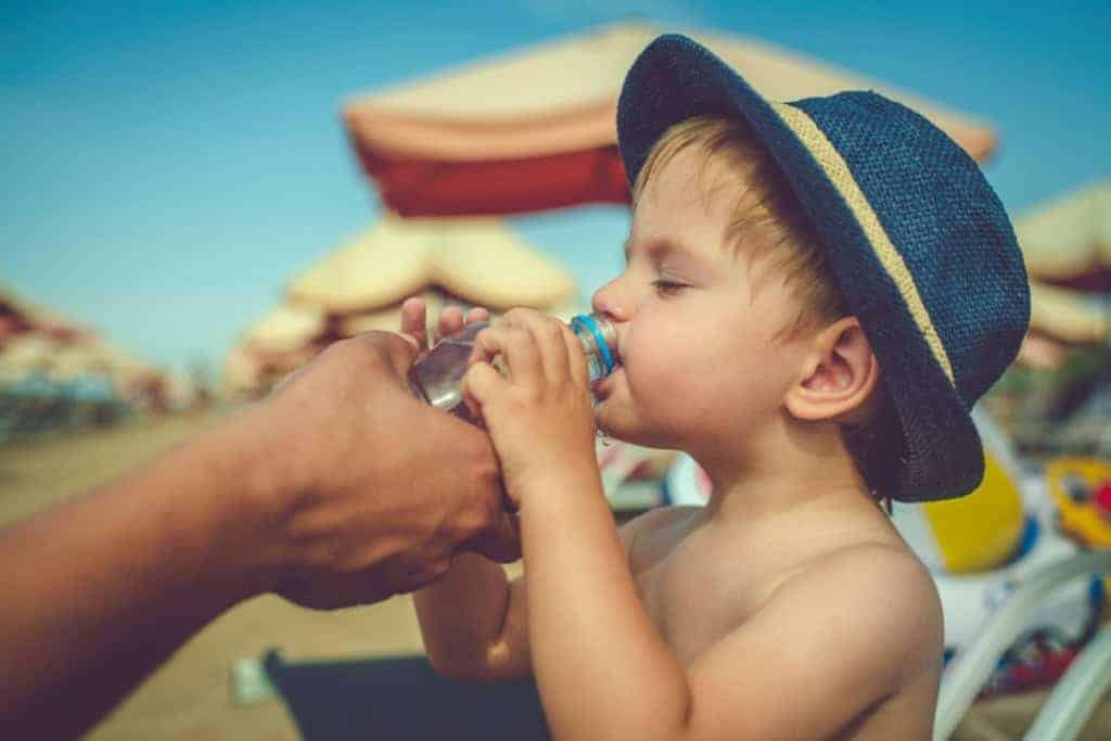 Young boy drinking water at the beach under an umbrella, representing international travel health coverage.