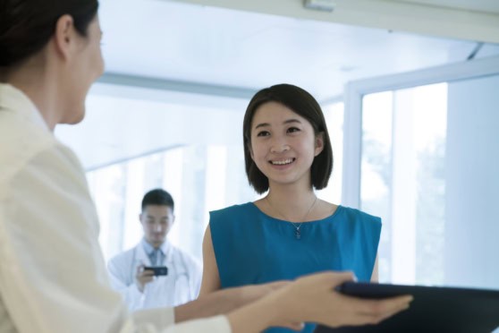 An Asian woman with a cheerful smile receiving assistance at a medical facility, illustrating comprehensive international health coverage for global travelers and expatriates.