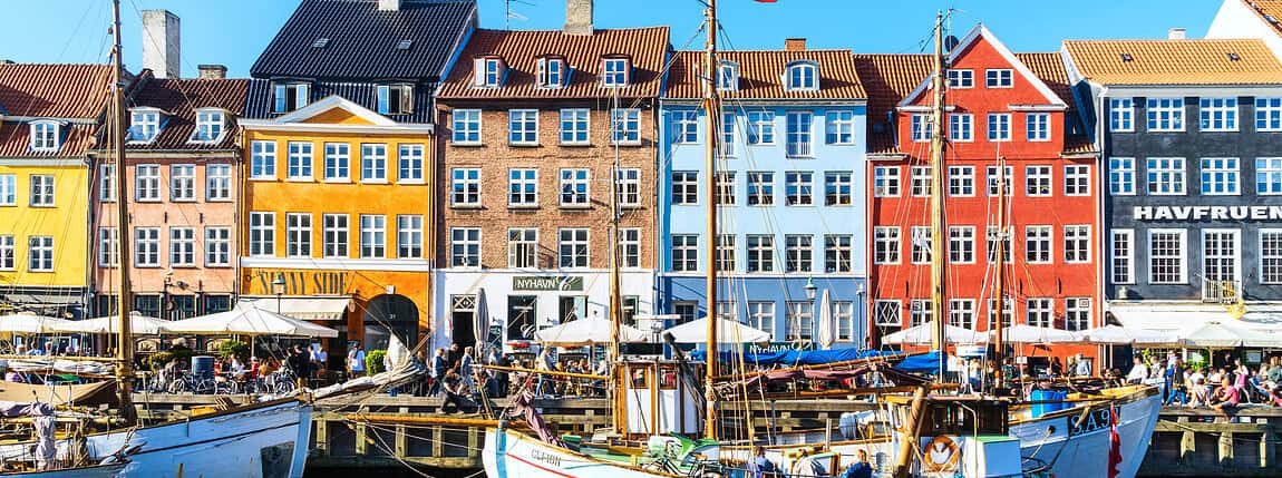 Colorful waterfront buildings in Nyhavn district, Copenhagen, Denmark, with boats docked along the harbor, vibrant atmosphere, and clear blue sky, showcasing Scandinavian architecture and lively harbor life.