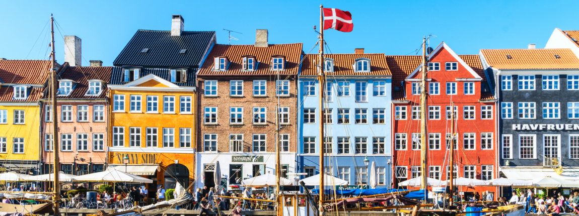 Colorful waterfront buildings in Nyhavn district, Copenhagen, Denmark, with boats docked along the harbor, vibrant atmosphere, and clear blue sky, showcasing Scandinavian architecture and lively harbor life.
