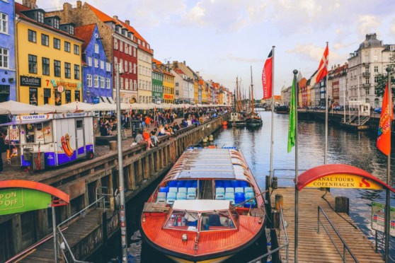 A bustling canal scene in Copenhagen with brightly colored houses, boats, and a lively promenade filled with visitors enjoying the waterfront ambiance.