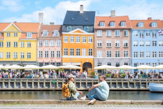 A picturesque European waterfront scene featuring multi-colored buildings, outdoor cafes, and tourists relaxing by the water’s edge.