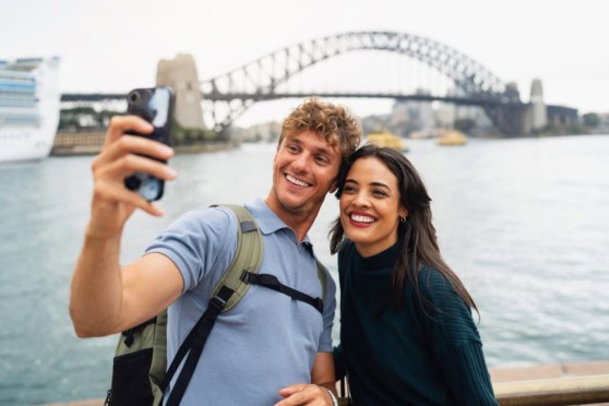 A happy couple enjoying sightseeing and capturing memories in Sydney, Australia by taking photos with the iconic bridge and harbor in the background.