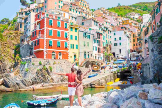 Bright, lively seaside scene featuring a picturesque Italian village with multicolored buildings, boats, and a couple exploring the harbor. Ideal for promoting international health insurance for travelers to Italy and scenic destinations.