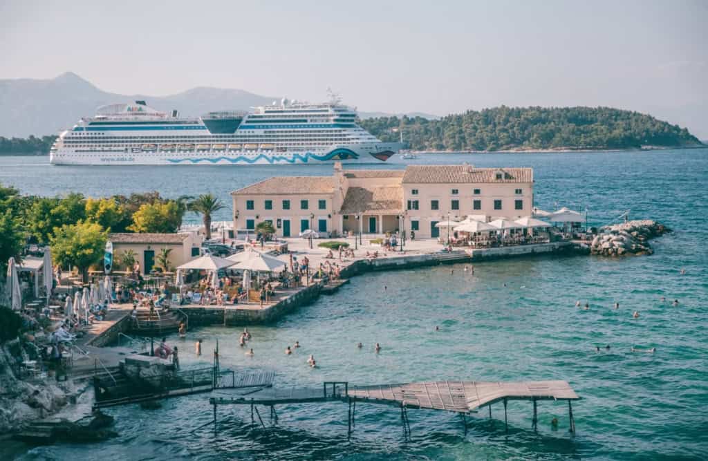 A large cruise ship sailing near a scenic coastal area with a lively waterfront, beachgoers swimming, and a charming seaside building with outdoor dining. A perfect destination for vacationers seeking relaxation and adventure.