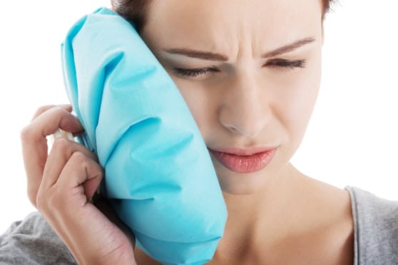 Close-up of a woman with a pained expression, pressing an ice pack to her ear to soothe discomfort.