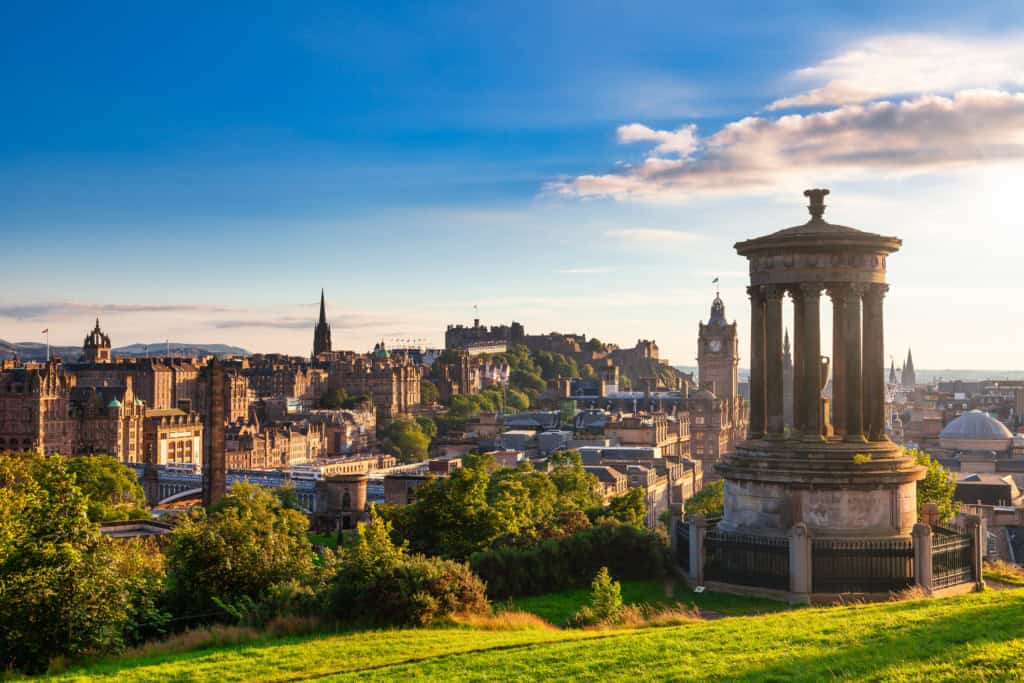 Bright sunny view of Edinburgh city skyline from Calton Hill, featuring historic monuments, Edinburgh Castle, and the clock tower with a clear blue sky and vibrant greenery.