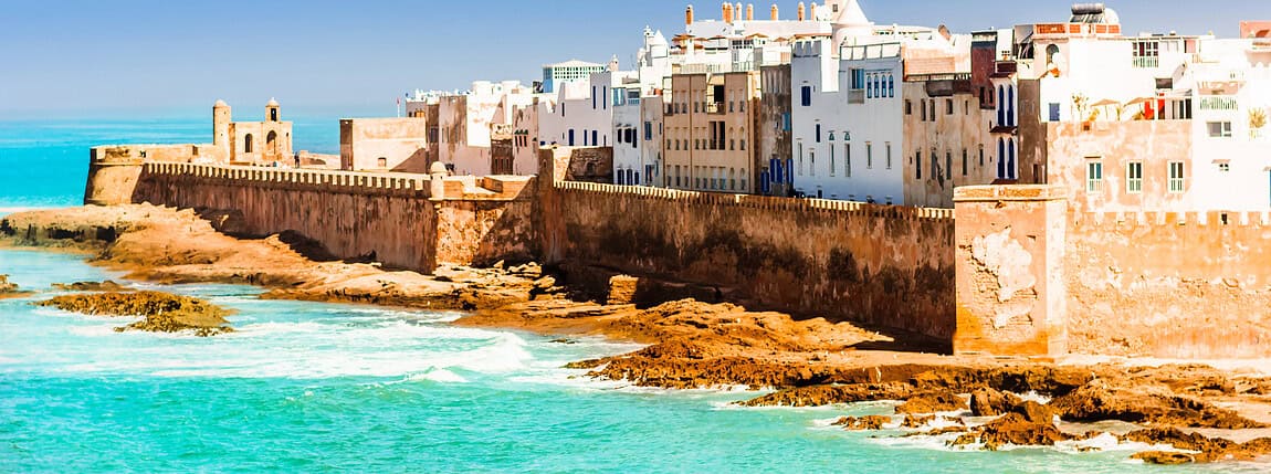 A panoramic view of a historic Moroccan city by the Atlantic Ocean, featuring white-washed buildings, a coastal fortress, and clear blue skies, highlighting travel and international living.