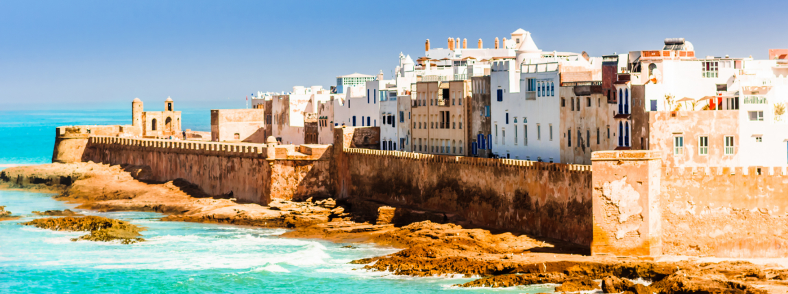 A panoramic view of a historic Moroccan city by the Atlantic Ocean, featuring white-washed buildings, a coastal fortress, and clear blue skies, highlighting travel and international living.