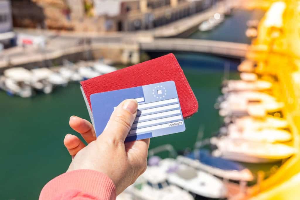 Image showing a person holding a European health insurance card with a marina and boats in the background, symbolizing international travel and healthcare coverage.