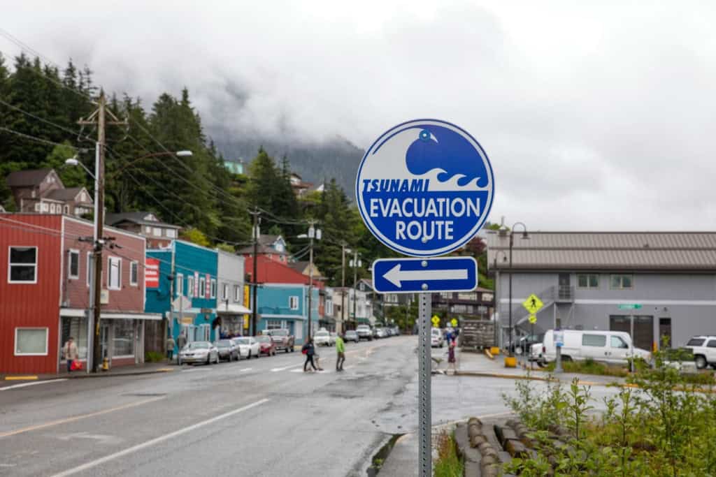 An image showing a blue tsunami evacuation route sign with an arrow pointing left in a seaside town, surrounded by colorful houses, parked cars, and a foggy mountain backdrop.