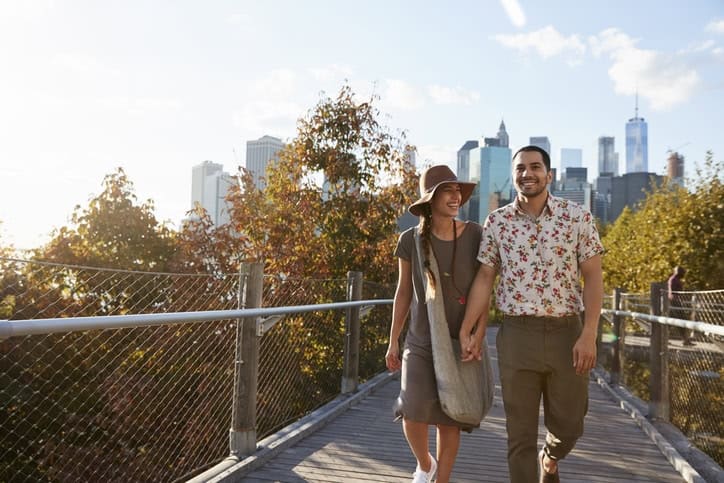 A joyful couple walking hand-in-hand on a wooden bridge in a city park with skyscrapers in the background, enjoying a sunny day with colorful fall foliage around. Ideal for illustrations of international travel, health insurance, and lifestyle.