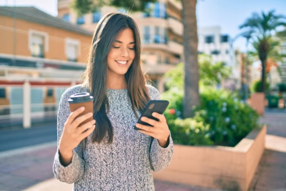 Woman smiling, holding coffee and phone outdoors, promoting international insurance services for travelers worldwide, with a sunny cityscape background.