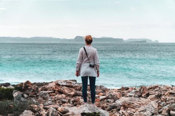 A woman with a camera on her neck gazing at the ocean from rocky shore, representing travel, exploration, and international insurance coverage for global adventures.