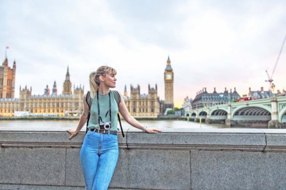 Image of a young woman traveler standing along the Thames River in London, with iconic Big Ben and historic houses of Parliament in the background, enjoying a scenic moment in the city.