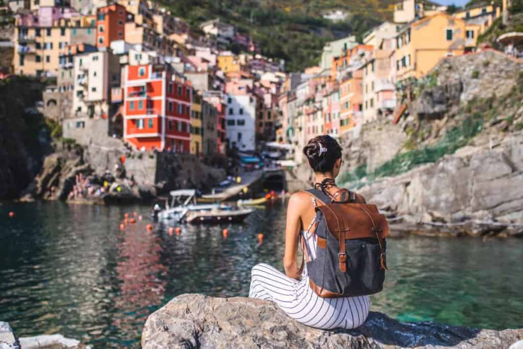 An international woman traveler with a backpack sitting on a rock, overlooking colorful hillside houses by the sea in a picturesque coastal town, promoting travel safety and insurance coverage.