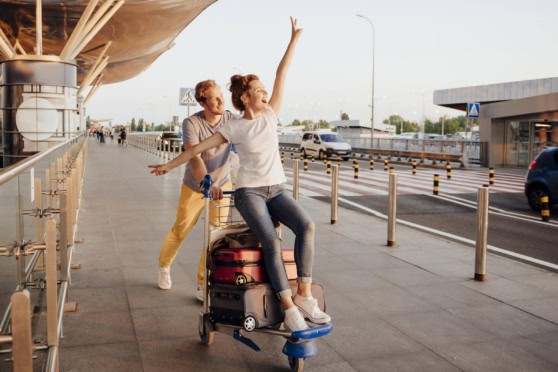 A happy couple at the airport, woman standing on luggage cart with arms raised, enjoying their travel experience with international insurance coverage.