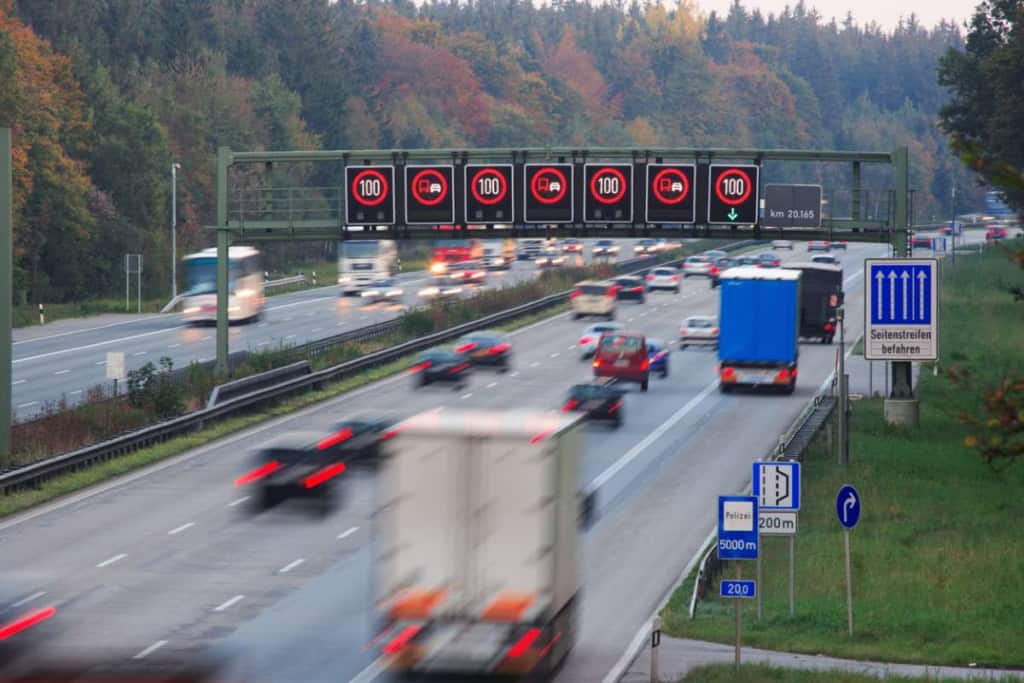 Digital highway sign showing 100 km/h speed limit and lane info, surrounded by busy traffic.