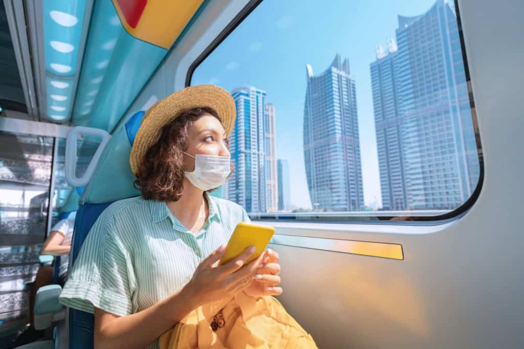 Woman in a protective medical mask rides the subway, holds a smartphone in her hands and looks out the window where the giant skyscrapers of Dubai rise
