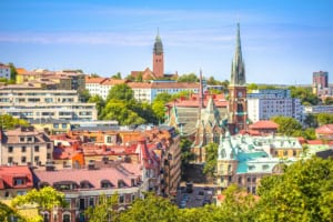 Nordic cityscape with historic church and modern buildings in bright daylight, featuring a blend of traditional and contemporary architecture, lush green trees, and a clear blue sky.