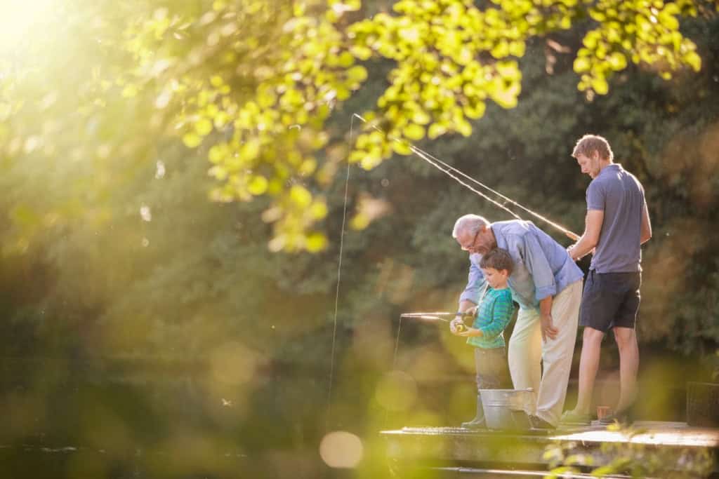 an eldery man sitting on a pier by a lake fishing with his two grandsons