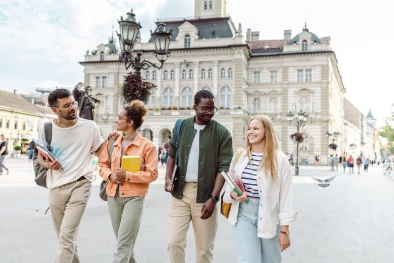 Alt text: Group of diverse young adults walking and talking in a city square, carrying books and backpacks, with historic buildings in the background.