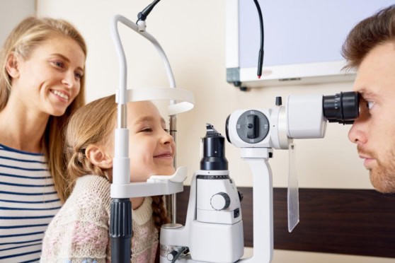 A young girl gets her eyes checked by an optometrist using a specialized slit lamp instrument while her mother watches with a smile. The setting is a modern eye care clinic dedicated to eye health screening.