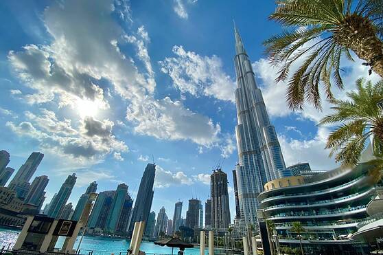 Modern Dubai skyline featuring the iconic Burj Khalifa, surrounded by skyscrapers, palm trees, and outdoor seating area along the waterfront, under a partly cloudy bright sky.