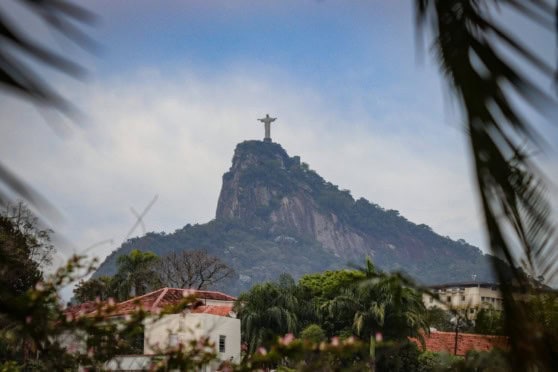 A majestic view of the Christ the Redeemer statue atop Corcovado Mountain in Rio de Janeiro, Brazil, with surrounding greenery and a partly cloudy sky. The image captures the iconic monument's grandeur, emphasizing travel safety and international insurance coverage for visitors and residents alike. The foreground includes tropical trees and rooftops, enhancing the scenic landscape of this famous tourist destination.