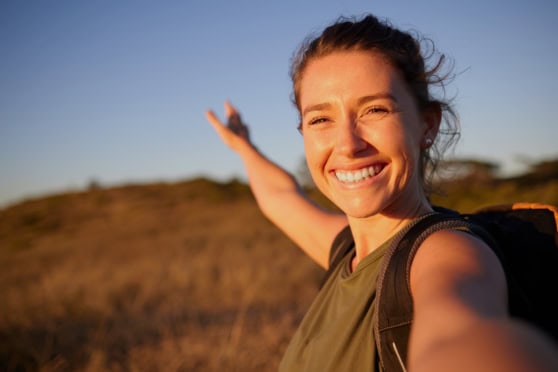 Happy woman with backpack taking selfie outdoors during day at sunset, illustrating international travel and health coverage for travelers worldwide.