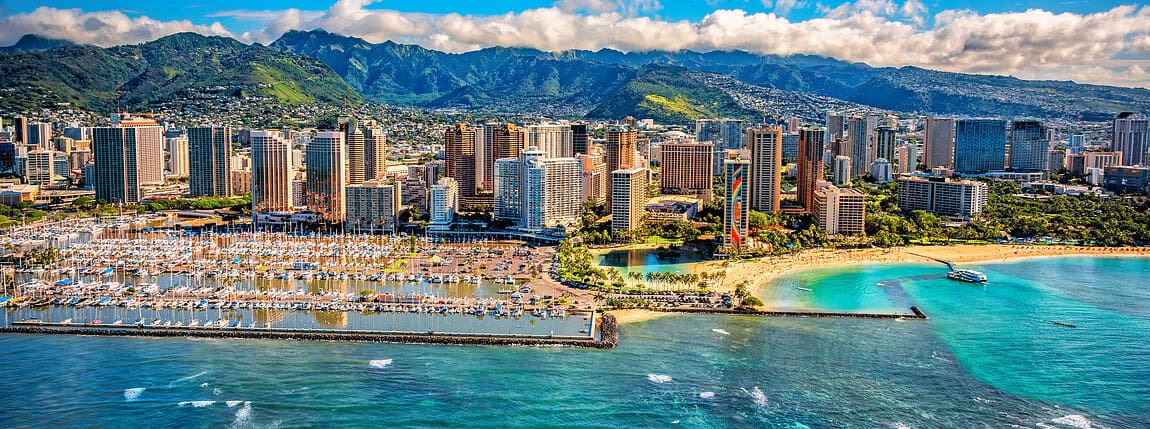 Downtown Honolulu skyline with high-rise buildings, marina, and turquoise ocean waters, set against lush green mountains on a sunny day.