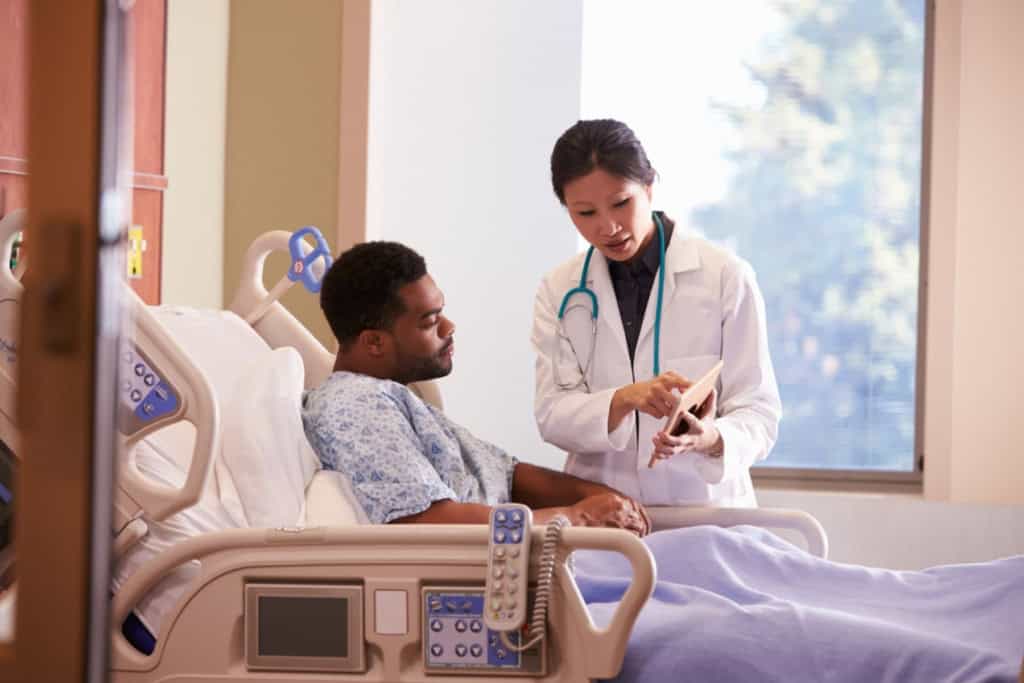 a male patient laying in a hospital bed talking to a female doctor, reflecting on the importance of having Schengen visa insurance when visiting Europe