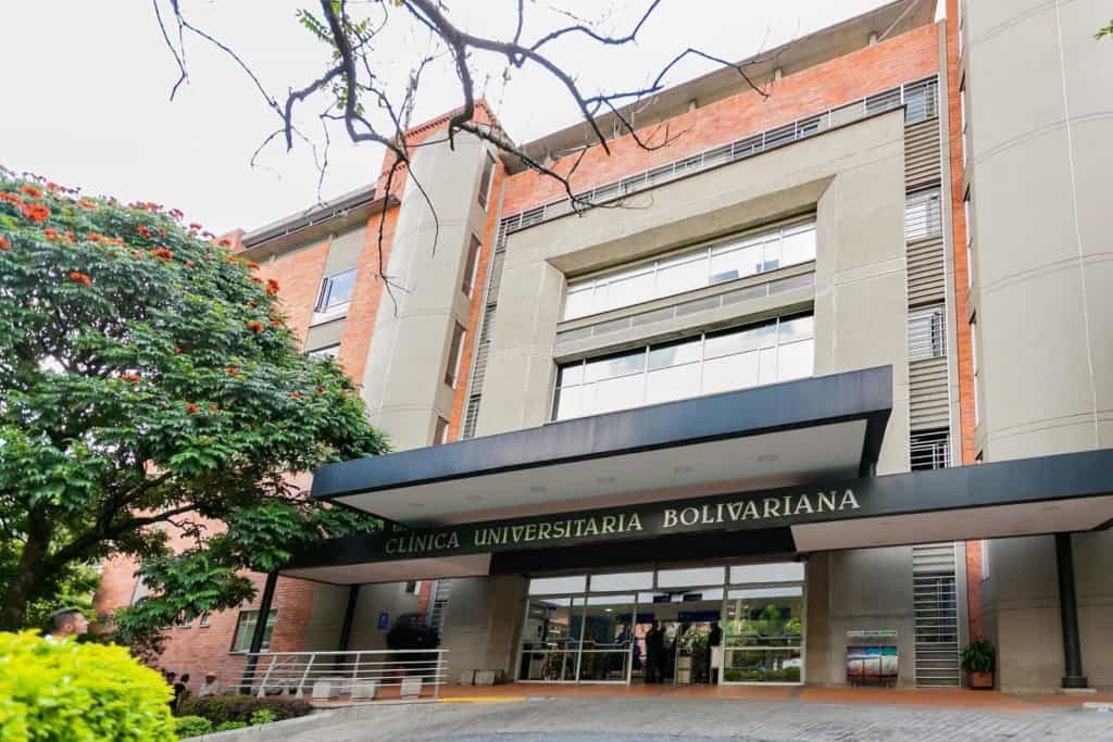Modern medical clinic building with a prominent entrance, lush greenery, and people outside, representing healthcare services in an urban setting.