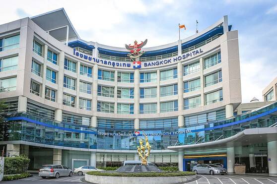 Modern multi-story hospital with glass facade, signage in Thai and English, and a decorative sculpture in the driveway area. Bright, sunny day with parked cars and a clear sky.