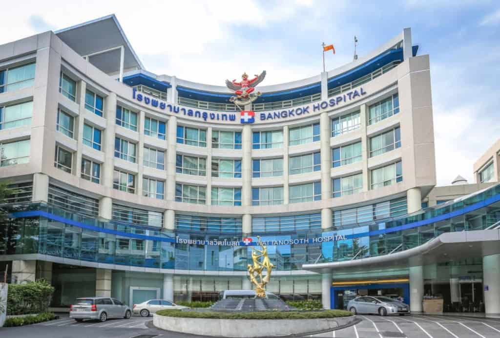 Modern hospital building with Bangkok Hospital and Wattanosoth Hospital signs, glass facade, parking lot, and a decorative golden sculpture in front.