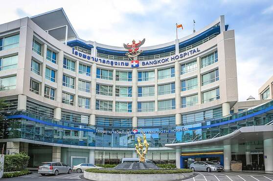 Modern hospital building with Bangkok Hospital and Wattanosoth Hospital signs, glass facade, parking lot, and a decorative golden sculpture in front.