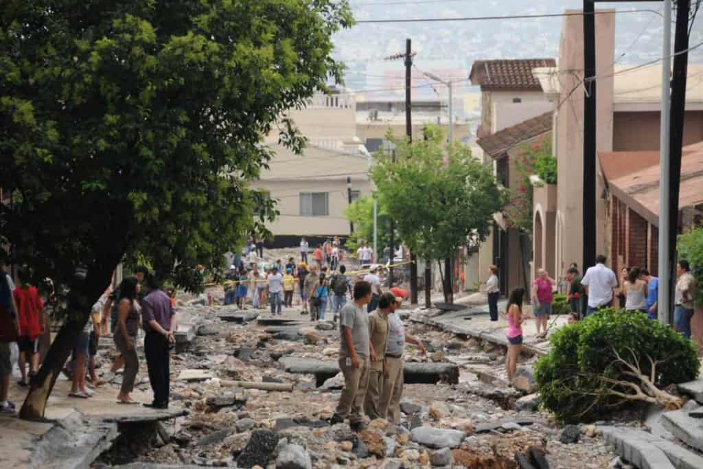 A street damaged by an earthquake with people inspecting the aftermath, fallen debris, and broken pavement in a city environment.
