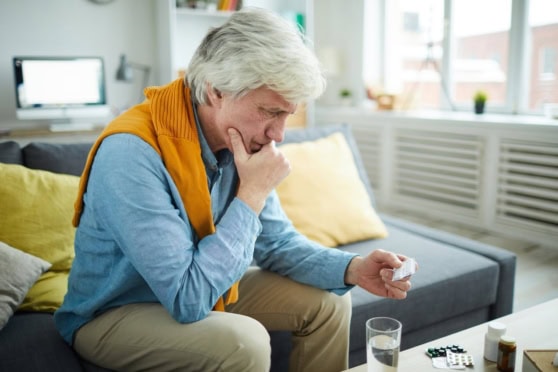 An elderly man sitting on a couch, holding medication and looking worried, in a bright and modern living room.