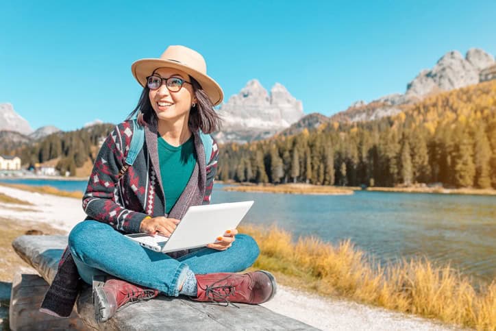 Experienced woman traveler with laptop by a scenic lake with mountains and trees in the background, emphasizing international health insurance coverage for expatriates and travelers worldwide.