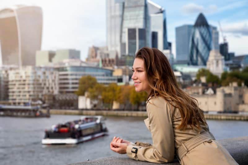 A smiling woman enjoying the view of London with the Thames River and city skyline in the background, highlighting the importance of international health coverage, including Americans moving abroad from the US.