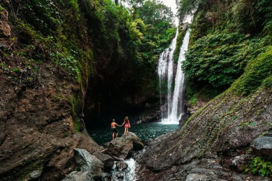 Scenic waterfall in dense forest backdrop featuring two explorers standing hand in hand near the water pool.