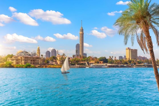 Alt Text: Scenic view of Cairo skyline with Nile River, sailboat, tall buildings including the Cairo Tower and palm trees, under a bright blue sky with clouds.