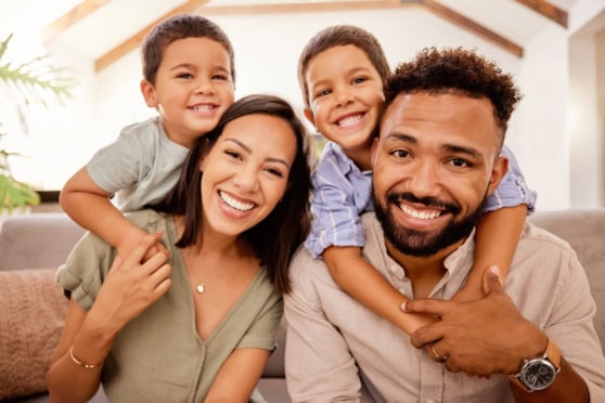 Happy multicultural family with two young boys on their parents' shoulders, smiling and embracing in a cozy living room setting, promoting health and life insurance coverage for international citizens.