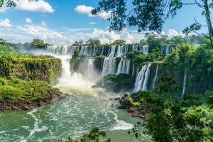 Vibrant cascade of Iguazu Falls surrounded by lush green rainforest under a bright blue sky with scattered clouds.