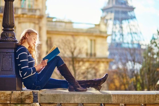 Woman reading a book outdoors, enjoying a sunny day in a park with historic architecture in the background.